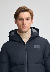 Puffjacka i mörk marinblå med en slät textur, med en framtill dragkedja, justerbar huva och en "POLO CLUB" logotyp på bröstet.