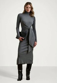 Γκρι μακρυμάνικο φόρεμα με ψηλό λαιμό, ντεκολτέ με κόμπο στη μέση και μήκος μέχρι το γόνατο. Συνδυάζεται με μια μαύρη υφασμάτινη τσάντα και μπότες μέχρι το γόνατο.