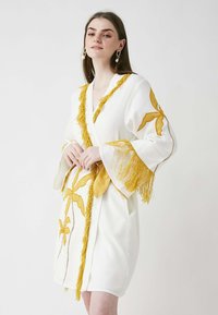 Balta halāta ar dzelteniem izšūtiem lapu rakstiem un frizētām akcentiem. Ietinama stila ar platām piedurknēm, papildināta ar atbilstošu jostu.