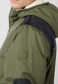 Chaqueta verde aceituna con detalles en negro, cuello forrado de forro polar suave y un parche de logo circular de goma en la manga. Textura suave.