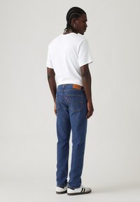 Hombre con cabello trenzado que lleva una camiseta blanca, jeans azules y zapatillas blancas con rayas negras, de pie y mirando por encima del hombro.