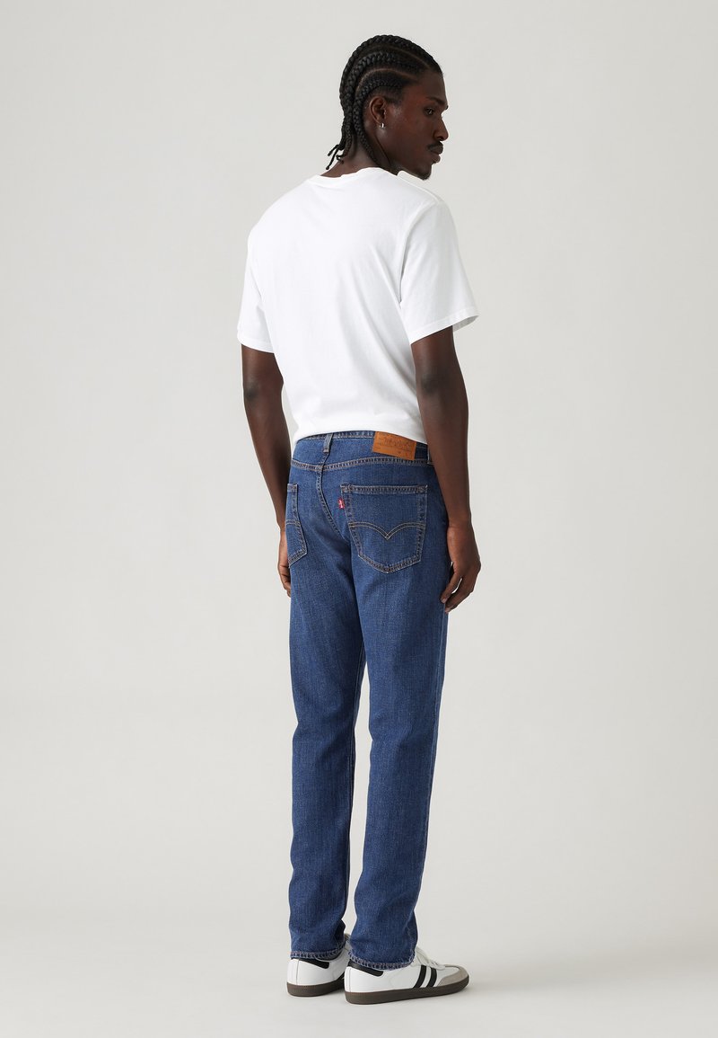 Mann mit geflochtenem Haar, der ein weißes T-Shirt, blaue Jeans und weiße Sneaker mit schwarzen Streifen trägt, steht und über die Schulter schaut.