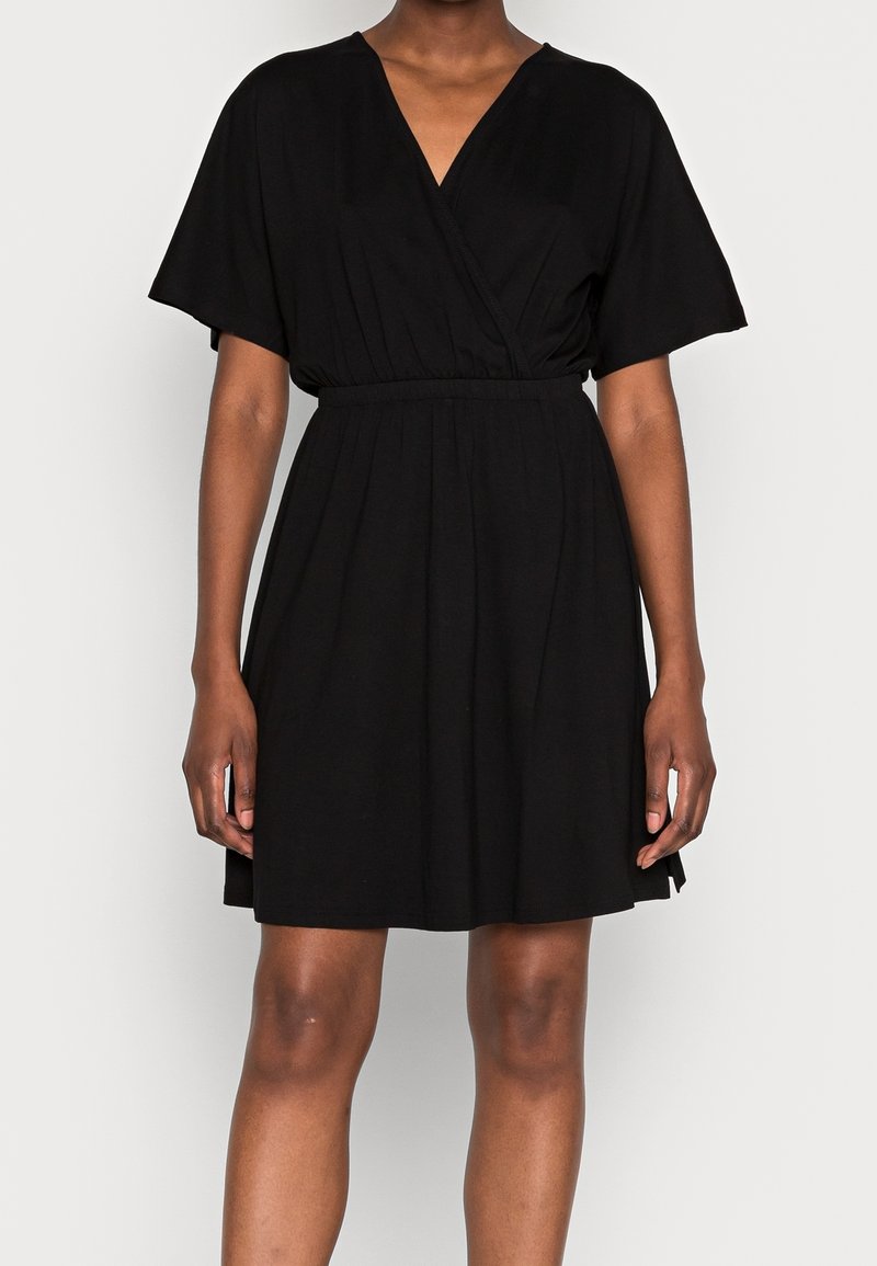 Femme portant une robe portefeuille noire courte avec des manches courtes et une taille élastique, debout devant un fond uni clair.