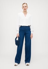 Weiße Bluse mit langen Ärmeln und V-Ausschnitt, kombiniert mit blauen, weiten Jeans mit goldenen Knöpfen und einer strukturierten maritimen Tasche.