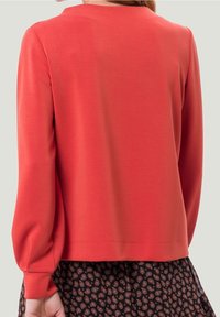 Czerwona bluzka z długim rękawem, okrągłym dekoltem i luźnym krojem. Gładka faktura, delikatne marszczenie przy rękawach, zestawiona z wzorzystą spódnicą.