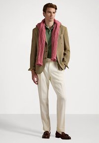 Νέος άντρας ντυμένος με μπεζ σακάκι, πράσινο πόλο, κρεμ παντελόνι, καφέ μοκασίνια και ροζ πουλόβερ περασμένο πάνω από τους ώμους, με φόντο λευκό.