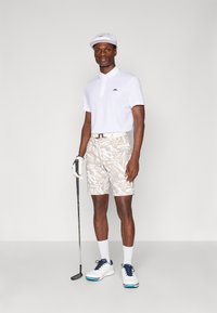 Vit polo tröja med svart logotyp, mönstrade beige shorts, vit keps, golfhandske och vita golfskor med marinblå detaljer. Står med en golfklubba.