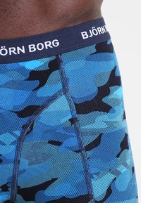 Sous-vêtements en camouflage bleu avec une ceinture élastique marine portant le texte "BJÖRN BORG." Le matériau semble lisse avec une finition légèrement texturée.