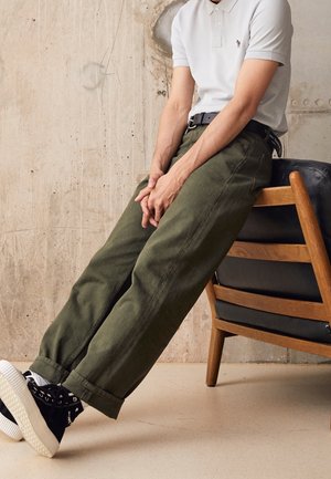 Personne portant un polo blanc, un pantalon vert olive et des baskets noires, assise sur une chaise en bois avec des coussins noirs, contre un mur en béton.