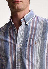 Hombre que lleva una camisa de botones a rayas azul, blanca y roja con cuello abotonado y un logo bordado en el bolsillo del pecho.