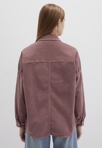 Übergrößiges Cordhemd in staubigem Rosé, mit klassischem Kragen, geknöpften Manschetten und abgerundetem Saum. Subtile gerippte Textur sichtbar.