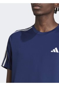 Sportovní tričko tmavě modré barvy z prodyšné tkaniny s bílými třípruhovými zdobeními na ramenou a malým logem na hrudi.