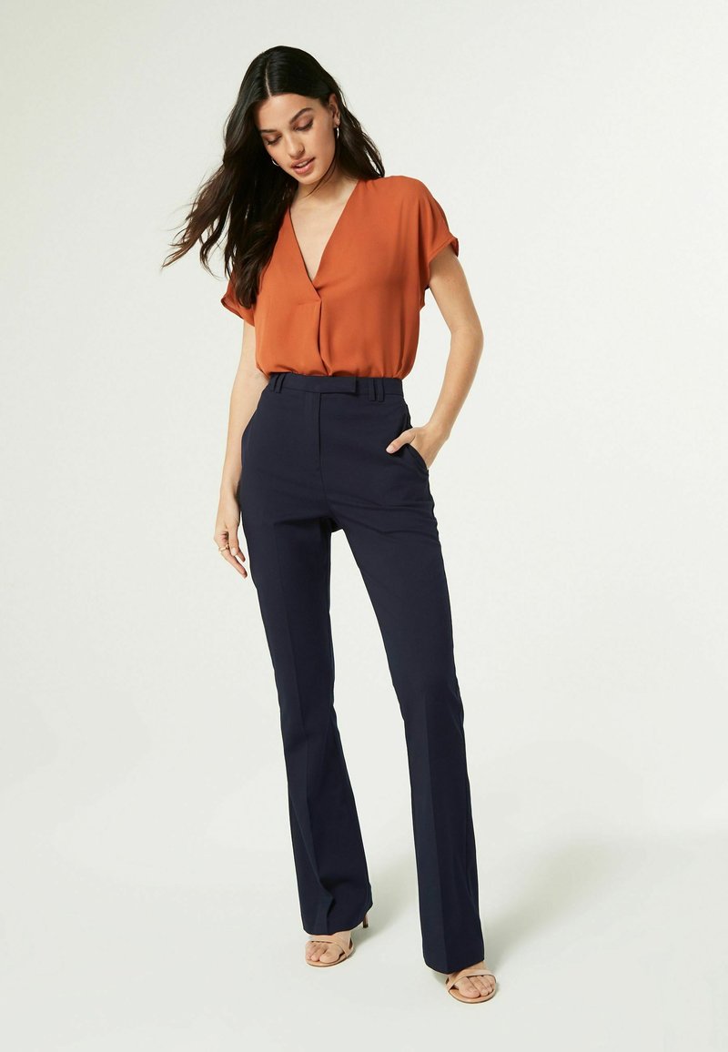 Roodachtige oranje blouse met een diepe V-hals en korte mouwen, gecombineerd met strakke, donkerblauwe uitlopende broeken. Glad materiaal en minimale details.