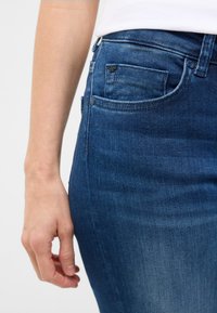 Jeans din denim albastru cu o textură netedă, având un buzunar frontal și un design mulat, cusături vizibile și un efect subtil de decolorare.