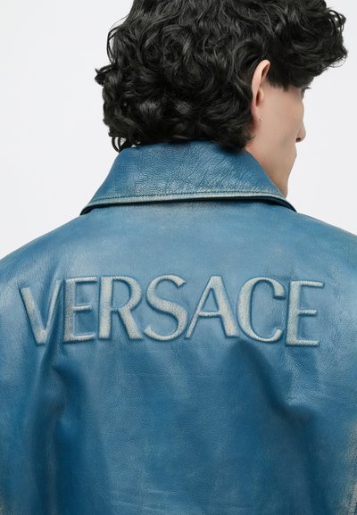 Άτομο με σκούρα σγουρά μαλλιά, στραμμένο πλάτη, που φοράει μπλε δερμάτινο μπουφάν με ανάγλυφο κείμενο "VERSACE" στην περιοχή του πίσω γιακά.