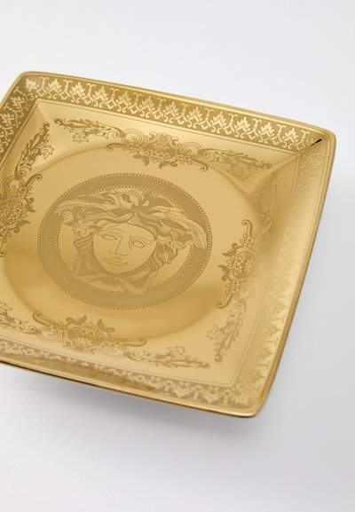 Τετράγωνος χρυσός δίσκος με περίτεχνα χαράγματα. Διαθέτει κεντρικό μοτίβο προσώπου περιβαλλόμενο από λουλουδένια σχέδια και περίτεχνες κορνίζες.