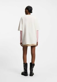 Oversized cremefarvet t-shirt lagdelt over en lyserød langærmet top, kombineret med en beige nederdel og sorte knæhøje støvler. Glat tekstur, afslappet pasform.