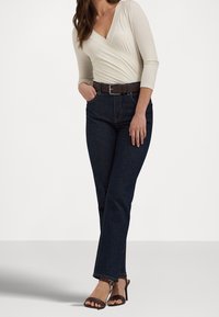 Cream long-sleeve wrap top with a v-neck, dark blue jeans with a straight leg, and brown belt. The model wears brown heeled sandals.