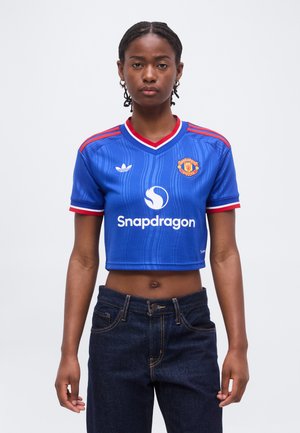 Jeune femme aux cheveux tressés porte un maillot de football Manchester United bleu court et un jean foncé, debout devant un fond uni.