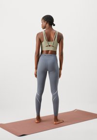 Oliven Grøn sports-bh med udsparingsdesign, parret med grå leggings med hvide accentlinjer. Modellen står barfodet på en yoga måtte.