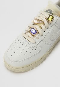 Zapatillas Nike Air blancas con cordones beige, decoradas con charms de joyas que incluyen una gema morada, una gema amarilla y un logo de Nike de strass.