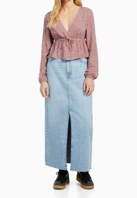 Blusa a maniche lunghe floreale rosa chiaro con vita arricciata, abbinata a una gonna maxi in denim azzurro chiaro con uno spacco frontale. Sneakers nere.