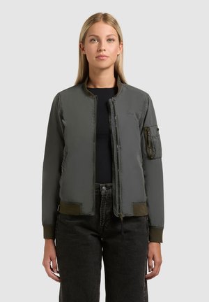 Femeie cu păr blond drept, purtând o geacă bomber verde închis peste un top negru și blugi negri, stând în fața unui fundal simplu.