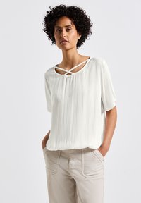 Weiße strukturierte Bluse mit rundem Ausschnitt und überkreuzten Trägerdetails. Kurze Ärmel und lockere Passform, kombiniert mit hellbeigen Hosen.