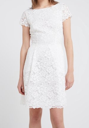 Kobieta ubrana w białą, krótką sukienkę z kwiatową koronką, dopasowaną talią i karbowanym dołem na jednolitym jasnym tle.