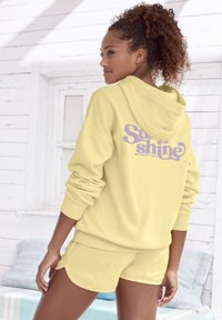 Žuta dukserica s kapuljačom i ljubičastim grafičkim detaljem "Sunshine" na leđima, u kombinaciji s odgovarajućim žutim šortsevima. Mekan, opušten kroj s rebrastim manžetama.