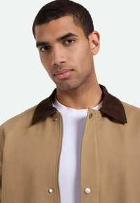 Beige Jacke mit einem braunen Cordkragen, ausgestattet mit Druckknopfverschlüssen und einem Reißverschluss vorne, getragen über einem weißen Hemd.