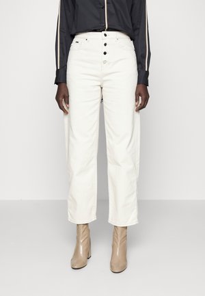 Persoon draagt witte jeans met hoge taille en zwarte knopen, een zwart shirt met lange mouwen en strepen op de mouwen, en beige enkellaarsjes.