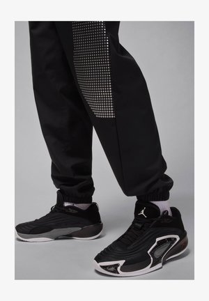 Tênis atléticos pretos com material texturizado, apresentando um design elegante, sola proeminente e detalhes brancos. Combinados com calças jogger pretas.