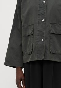Casaco verde escuro com mangas oversized, fecho frontal com botões de pressão e dois bolsos no peito. O tecido parece durável com uma textura suave.
