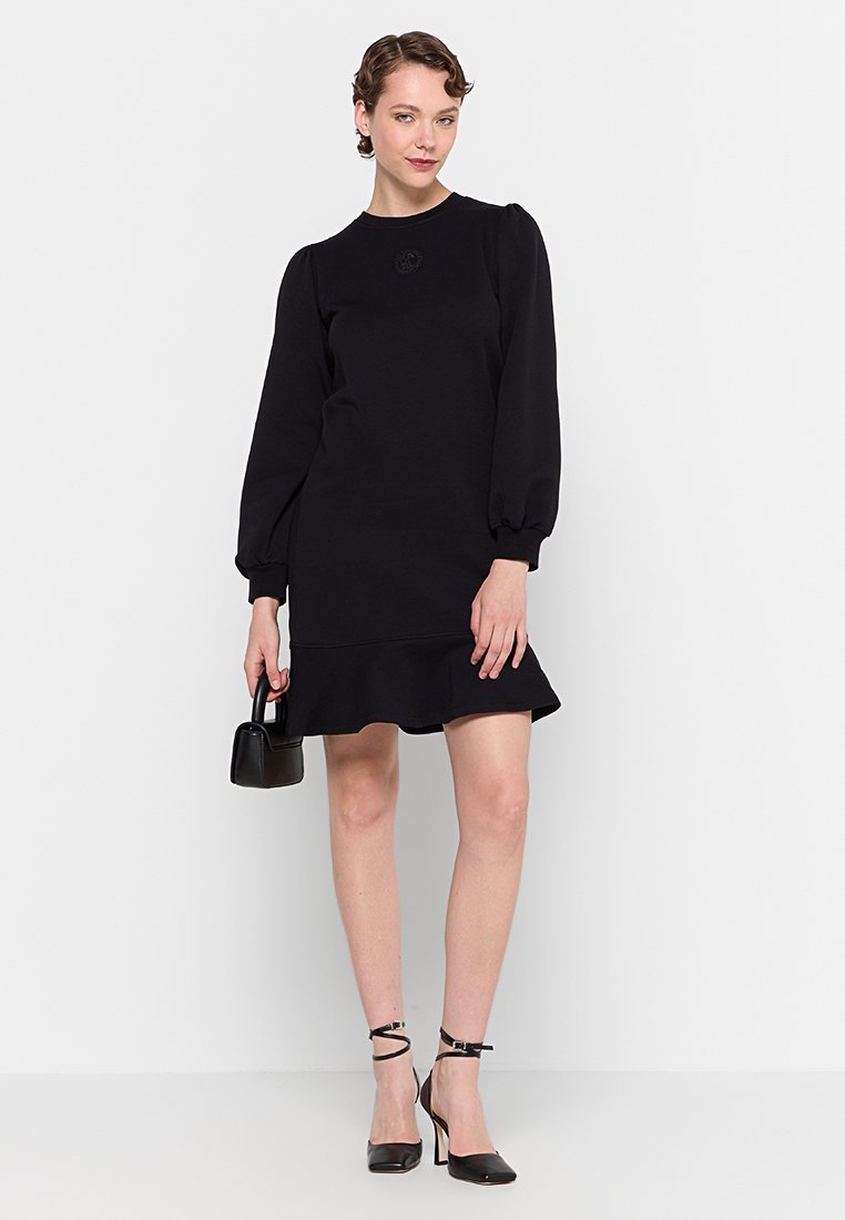 Robe noire avec de longues manches volantes, un col rond et un ourlet froncé. Texture lisse, design légèrement ajusté, accompagnée de talons noirs.