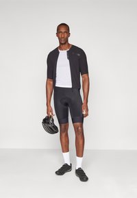 Tenue de cyclisme comprenant un maillot noir à manches courtes, une chemise intérieure en mesh blanche, un short rembourré noir et des chaussures de cyclisme noires, tenant un casque.