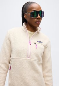 Γυναίκα που φοράει μεγάλα αντανακλαστικά γυαλιά ηλίου και ένα εκρού fleece της Patagonia με ροζ λεπτομέρειες στο φερμουάρ, κοιτάζοντας λίγο προς τα δεξιά.