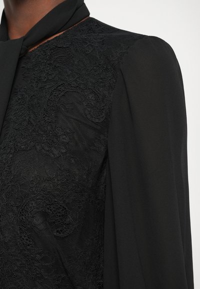 Melnā blūze ar teksturētu mežģīņu dizainu vienā pusē, gludu audumu otrā pusē, ar sasietu kakla detaļu un garām piedurknēm.