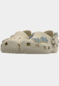 Sabots beige à enfiler avec des breloques en forme de fleurs bleues et des trous de ventilation sur les orteils et les côtés, vus en léger contre-plongée.