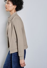 Giacca beige chiaro con design aderente, chiusura con zip e tasche laterali. Polsini e orlo a costine, realizzata in un tessuto liscio.