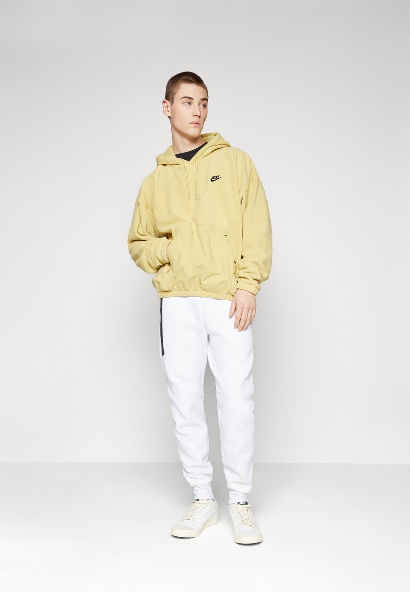 Sudadera amarilla con capucha y un logo negro, combinada con pantalones de chándal blancos y zapatillas blancas. Tela texturizada, puños y cinturilla elásticos.