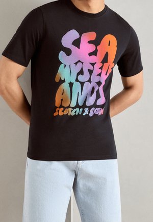 Mężczyzna w czarnej koszulce z kolorowym napisem "Sea Myself And I Scotch & Soda", w jasnoniebieskich dżinsach na jednolitym tle.