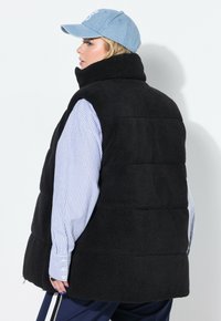 Svart fleecemidja med hög krage, med vertikal sömdetaljering och strukturerad yta, lager på lager över en blå och vit randig skjorta.
