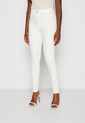 Femme portant un jean skinny blanc taille haute et des sandales blanches à talons hauts ouvertes à l’avant, debout devant un fond blanc uni.