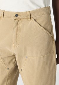 Khaki cargo pants made of durable cotton, featuring multiple angled pockets, visible stitching, and a metal button closure at the waist.