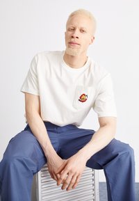 Witte katoenen t-shirt met een borstzak met een geborduurd ontwerp. De persoon draagt een marineblauwe broek en zit op een zilveren oppervlak.