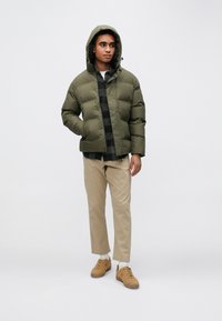 Olívazöld steppelt dzseki kapucnival, fekete és szürke kockás ing fölött viselve, világos bézs nadrággal és világosbarna tornacipővel kombinálva.