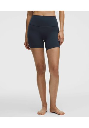 Kvinde iført mørkeblå fitness-shorts med høj talje, stående barfodet mod en ensartet lys baggrund.