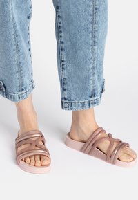 Pink slide sandals with intertwining straps, smooth texture, and a thick, chunky sole. Worn with cuffed light blue jeans.