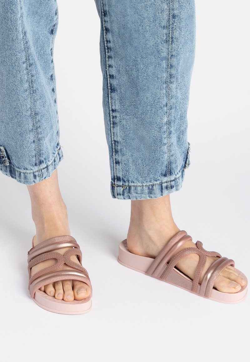 Pink slide sandals with intertwining straps, smooth texture, and a thick, chunky sole. Worn with cuffed light blue jeans.